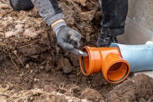 A person wearing gloves installs an orange PVC pipe with a T-joint underground, surrounded by dirt and soil, for what appears to be plumbing or drainage work.